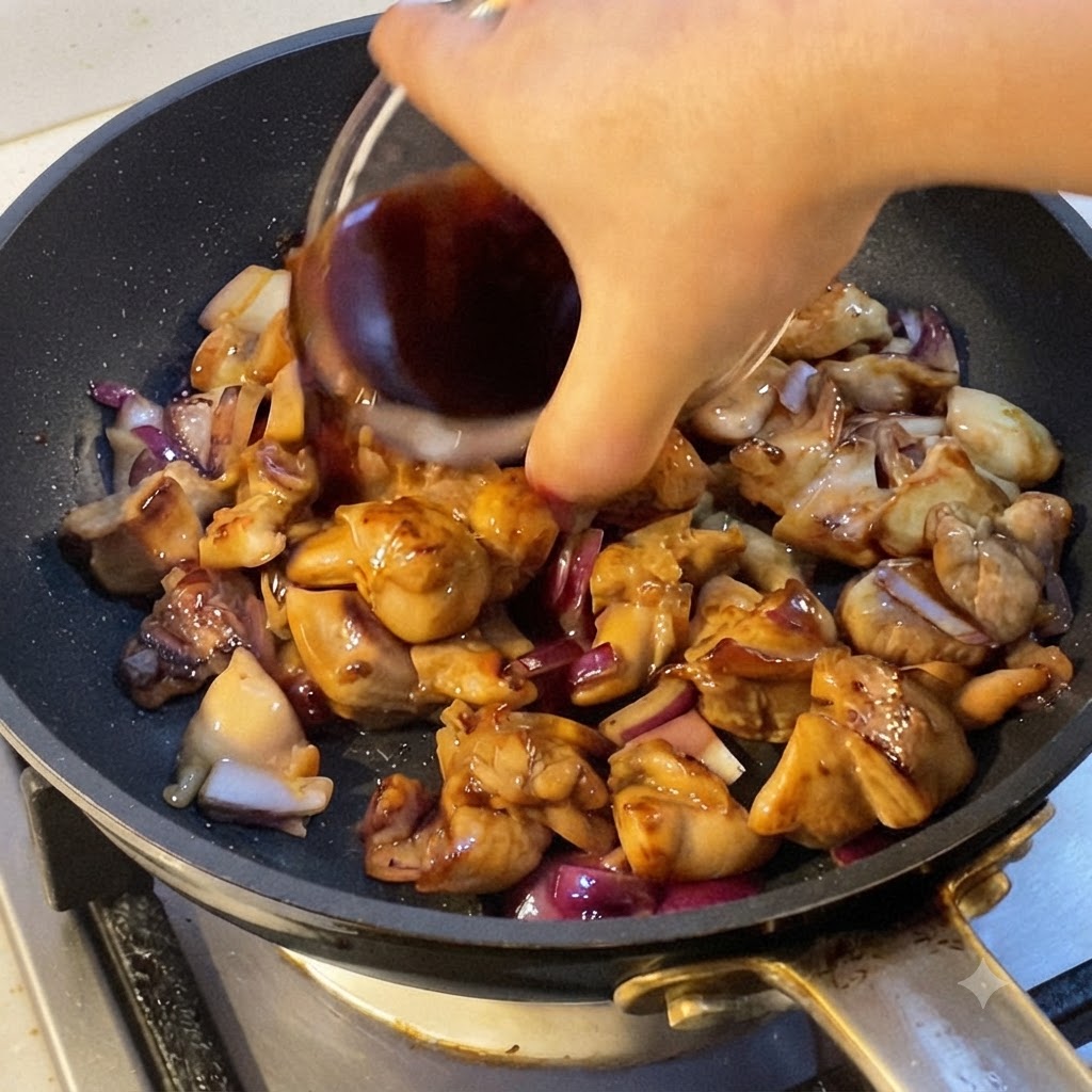 A hand pouring dark cooking sauce into a pan filled with cooked chicken pieces and red onion chunks.