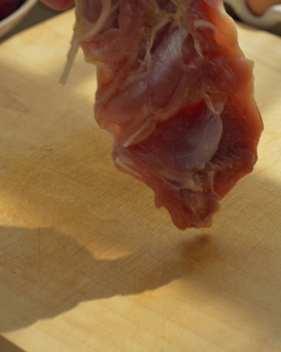 Boneless chicken thigh fillets being cut into large pieces on a wooden cutting board with a knife.