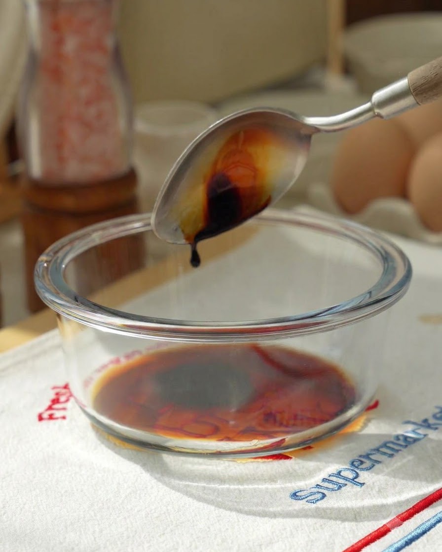 Soy sauce being poured from a spoon into a glass mixing bowl containing sauce ingredients.