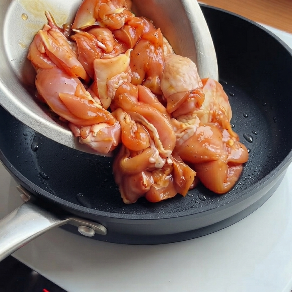 Marinated chicken pieces being poured from a bowl into a hot black frying pan.