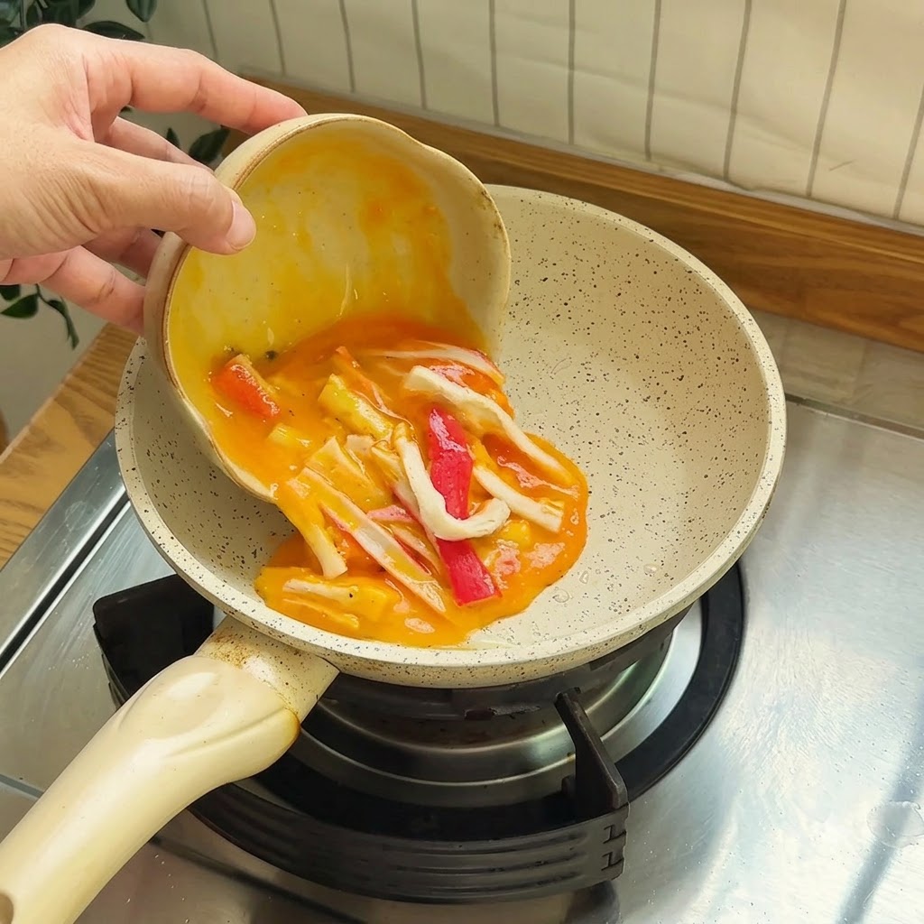 Beaten eggs mixed with red and white crab stick pieces being poured into a small non-stick pan.