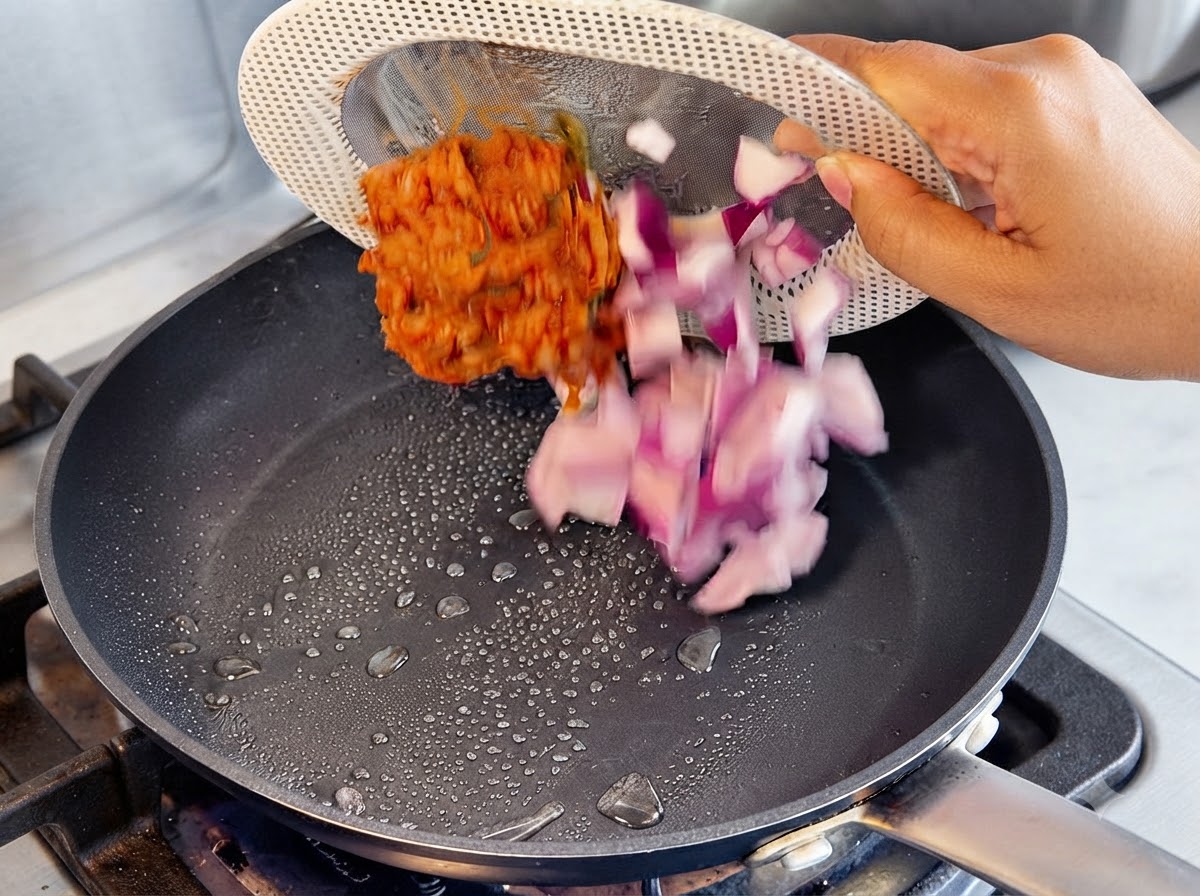 Chopped purple onions and red kimchi being poured from a strainer plate into a hot frying pan with oil droplets.
