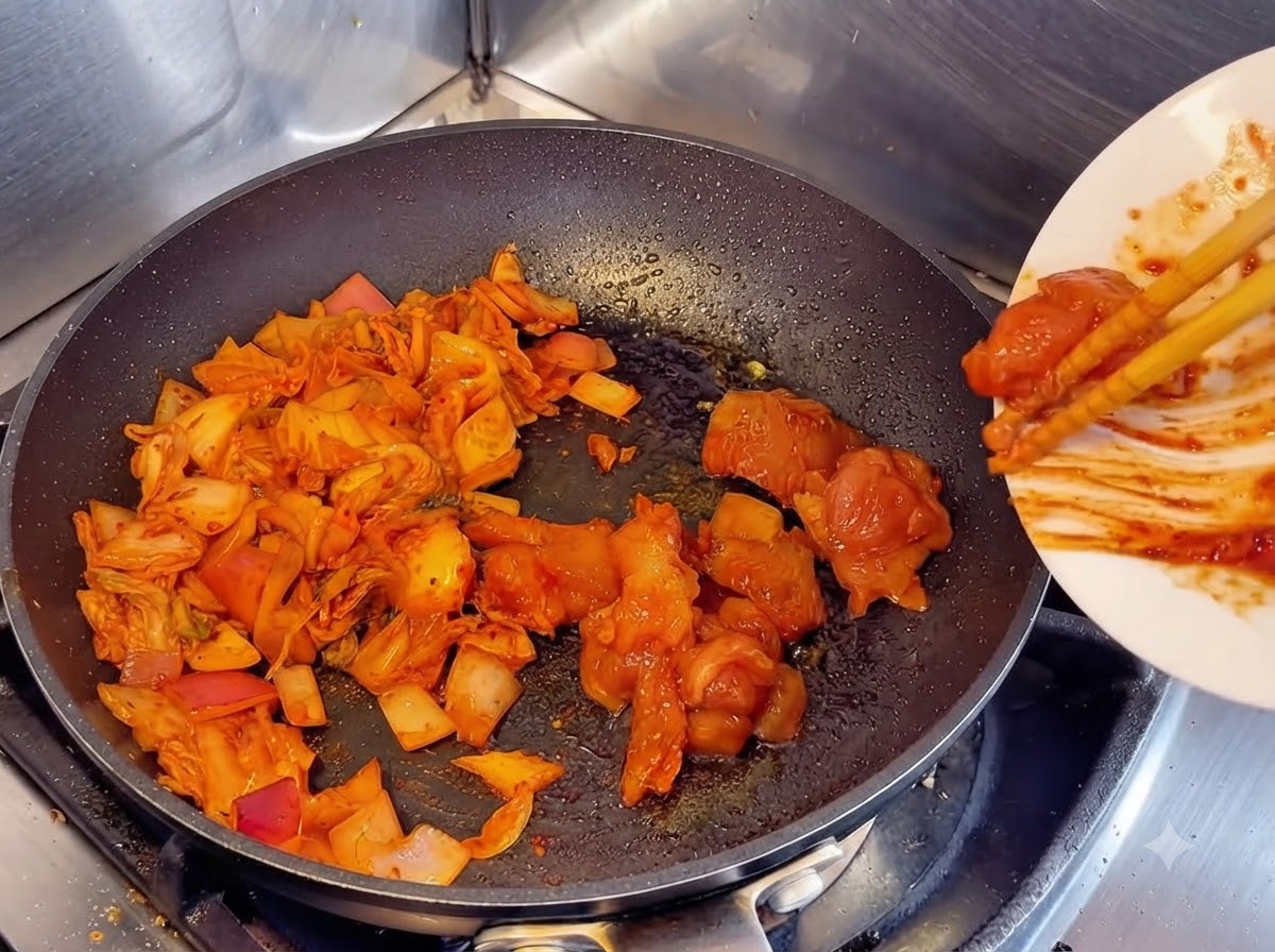 Using chopsticks to slide marinated raw chicken chunks from a white plate into a frying pan next to sautéed kimchi and onions.