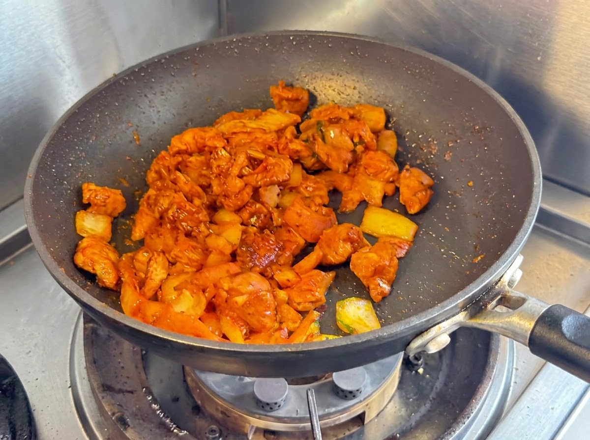 Diced chicken breast onions and chopped kimchi being stir fried together in a dark non stick pan.