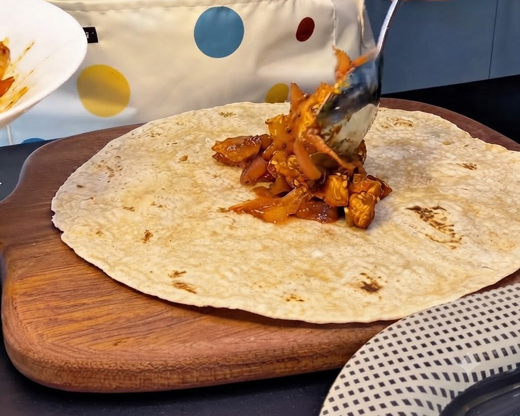 A large spoonful of spicy chicken and kimchi filling being placed onto the center of a whole wheat tortilla.