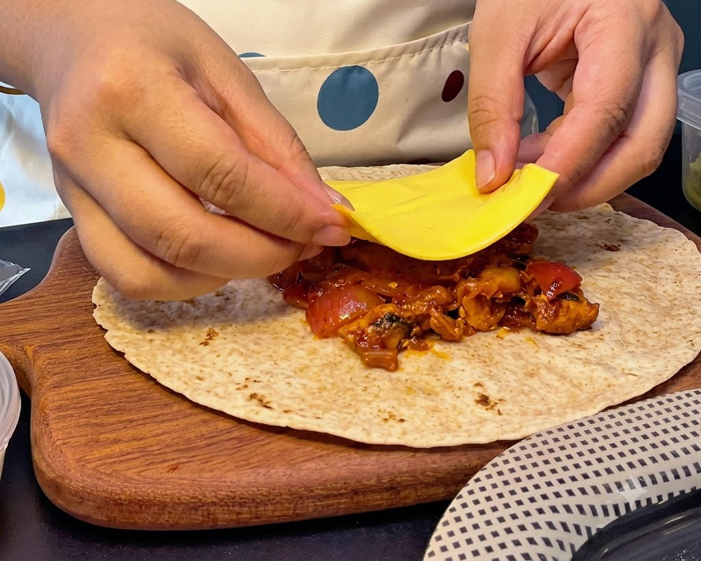 Hands placing a square slice of yellow cheese over a pile of hot chicken and kimchi filling on a flatbread.