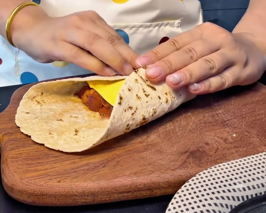 Hands folding the edges of a flatbread tightly over a slice of cheese and filling to create a sealed square packet.