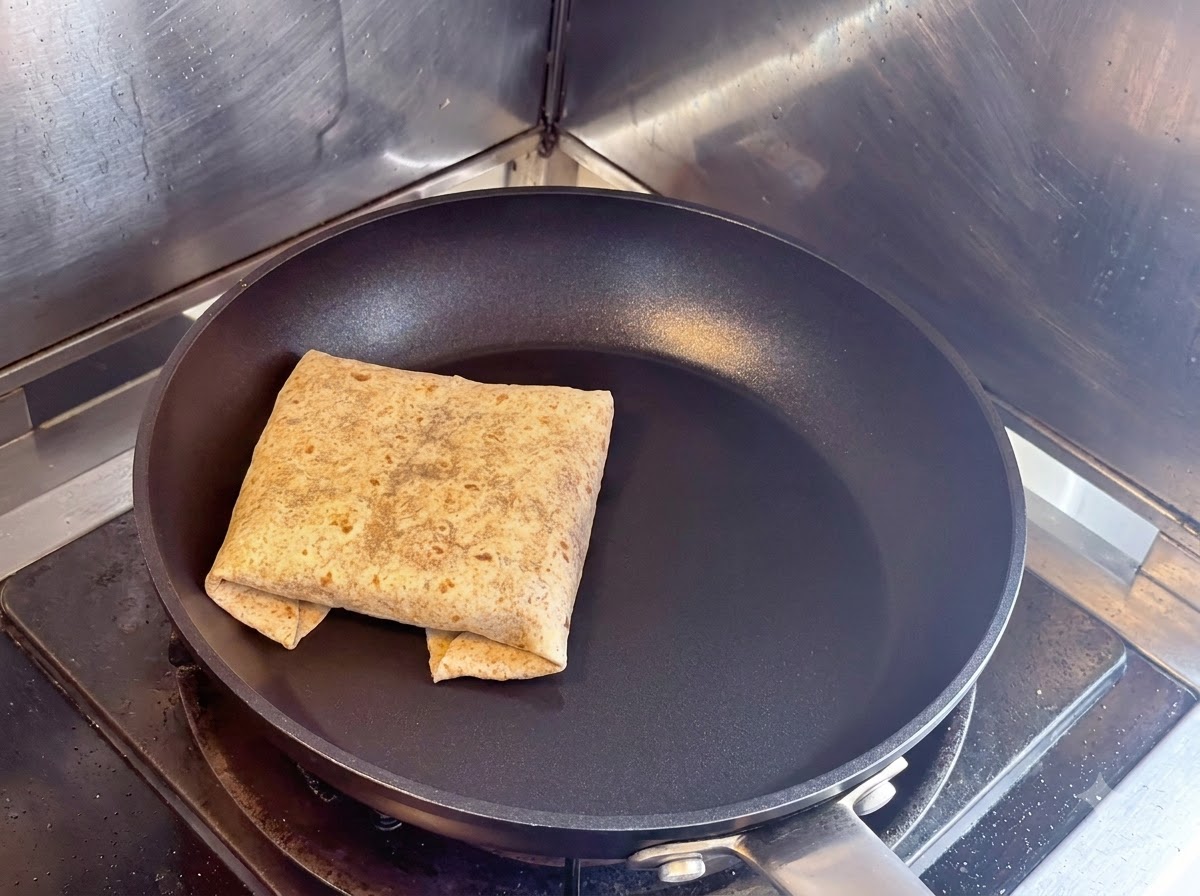A folded square flatbread wrap toasting seam-side down in a dry black non-stick skillet.
