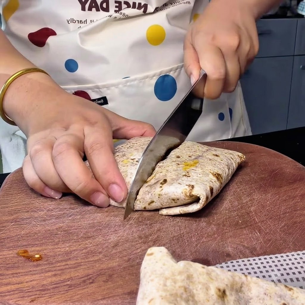 A person using a large chef knife to cut a diagonally arranged whole wheat wrap on a wooden cutting board.