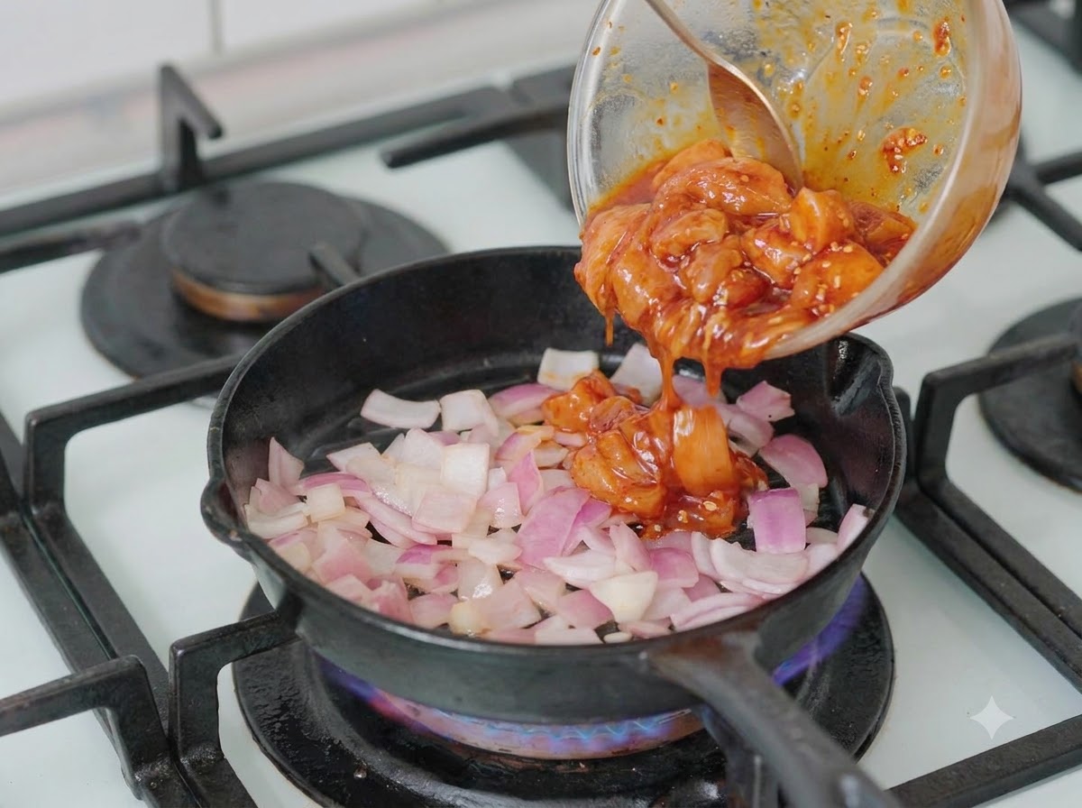 Marinated chicken pieces being poured from a bowl into a pan containing chopped onions.