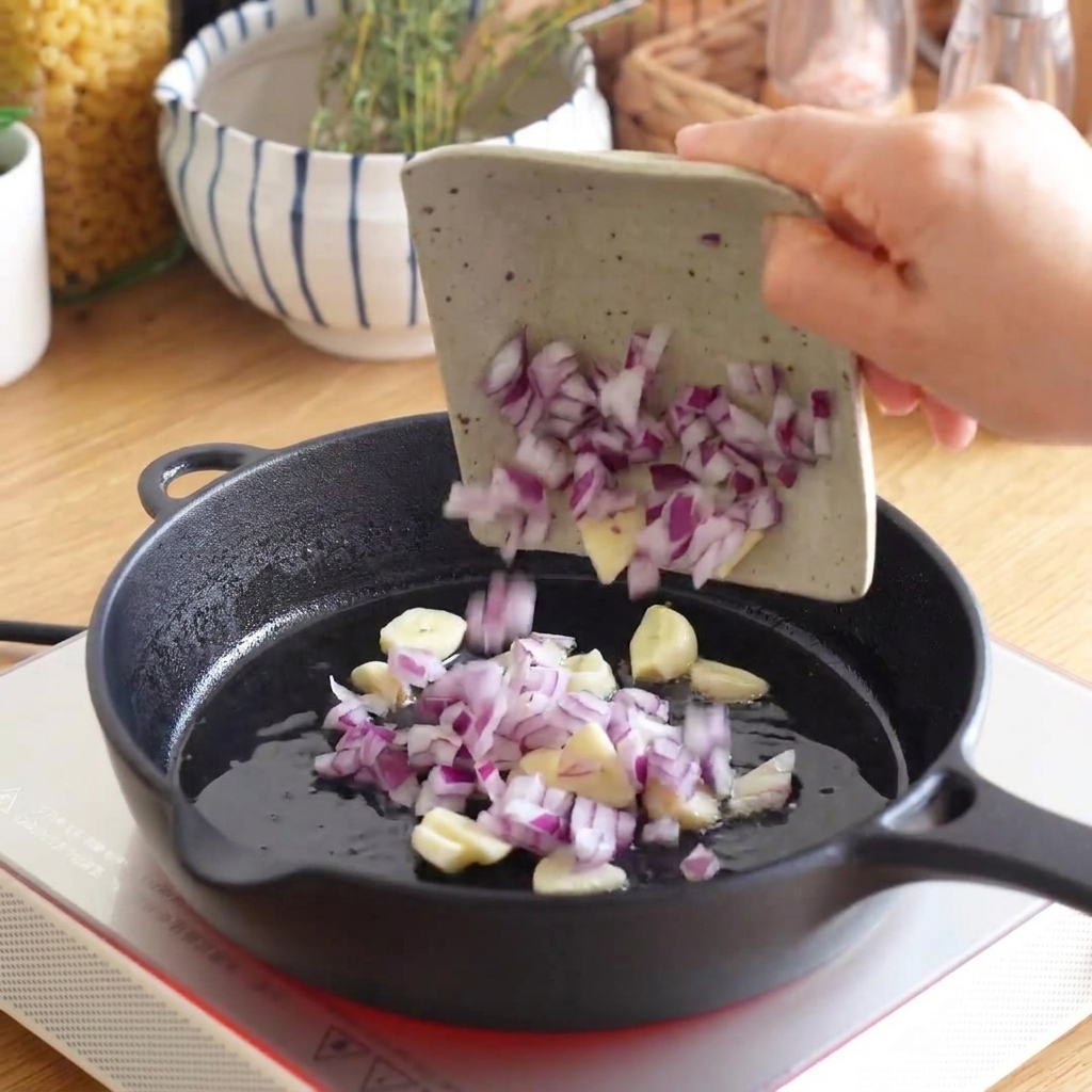 Chopped red onion and sliced garlic being poured from a small dish into a hot cast iron skillet.