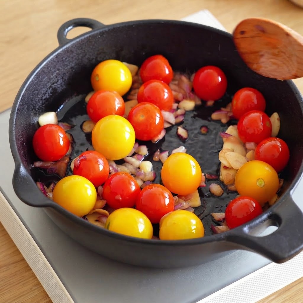A wooden spoon held above a skillet containing cherry tomatoes, chopped onions, and garlic.
