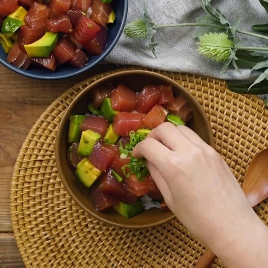 A hand sprinkling chopped green onions over the tuna and avocado bowl.