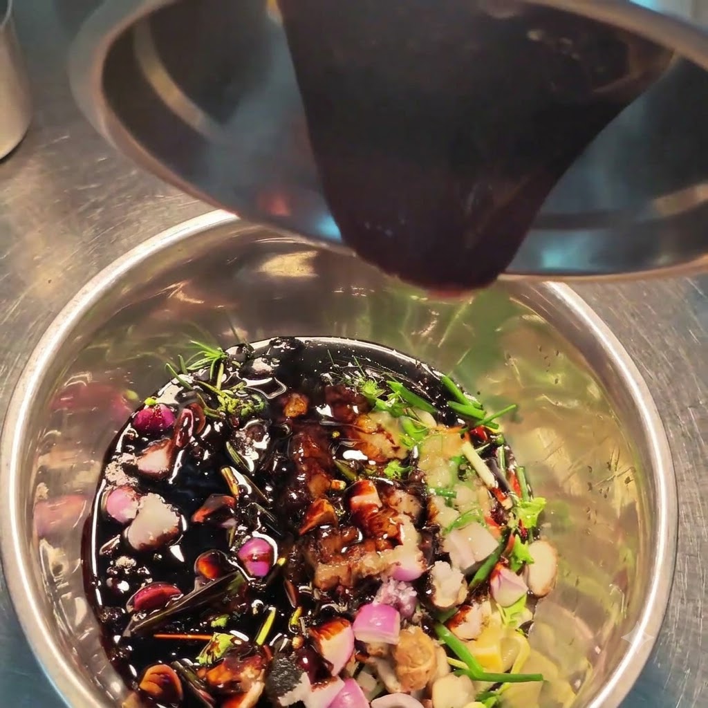 Dark sauce being poured from a steel container into a bowl filled with various herbs, spices, and chopped aromatics.