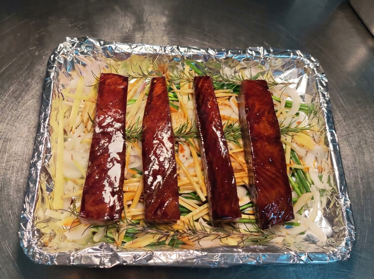 Four marinated tuna blocks neatly arranged in a row over a bed of shredded ginger, onions, scallions, and rosemary on a foil-lined baking sheet.