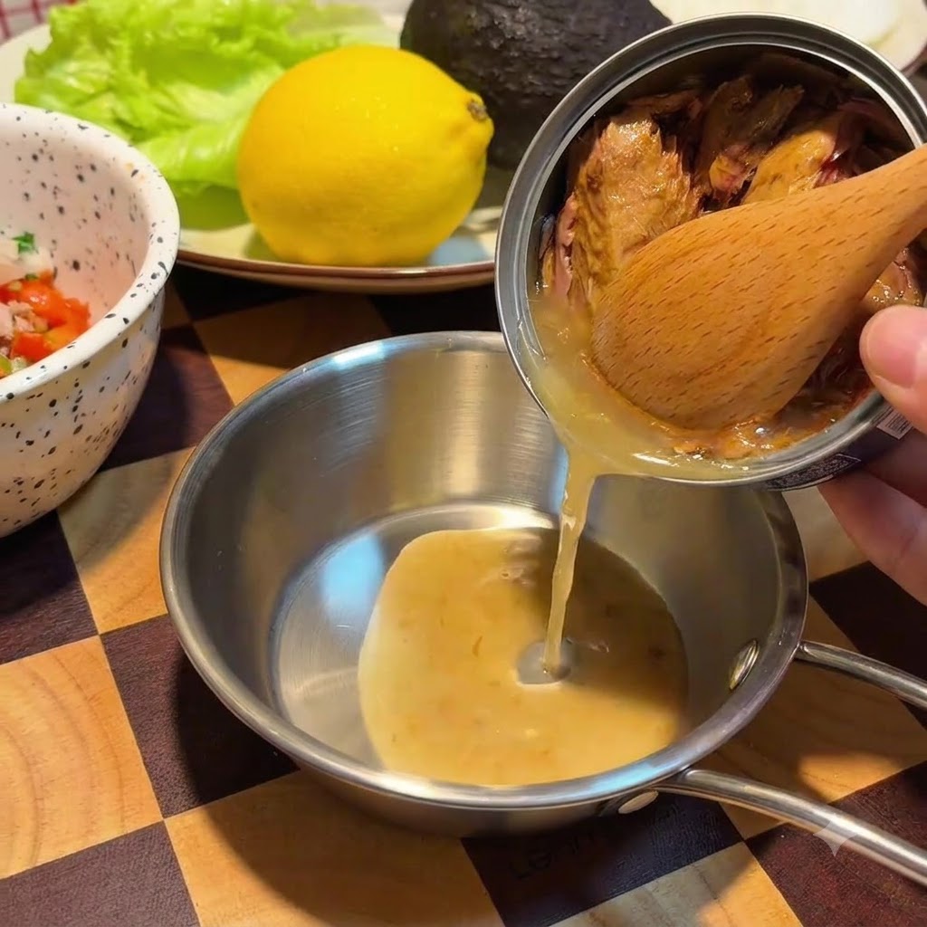 A wooden spoon holds back tuna chunks inside a metal can while golden oil pours into a small stainless steel bowl.