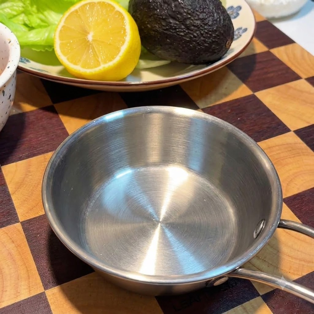 An empty small stainless steel bowl sitting on a checkered wooden board next to a plate with a lemon and an avocado.