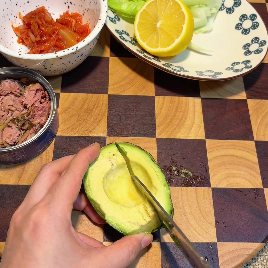 A hand using a knife to slice checkered cubes into an avocado half resting on a checkered wooden cutting board.