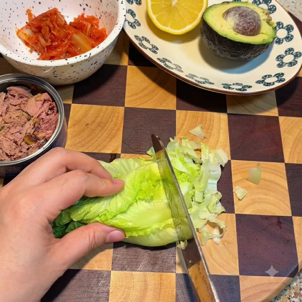 A knife slicing through fresh green lettuce leaves on a checkered cutting board next to a bowl of kimchi and a canned tuna.