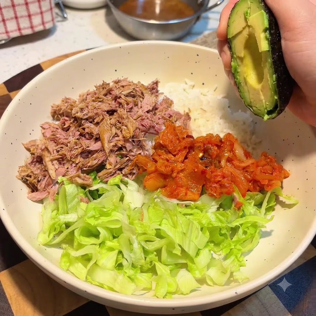 A hand holding an avocado half over a large bowl containing rice, lettuce, tuna, and kimchi.