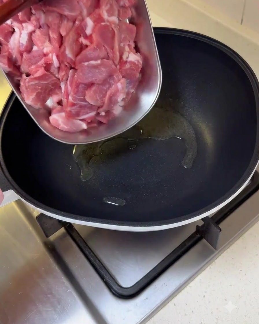 Sliced raw pork pieces being transferred from a tray into an oiled frying pan.