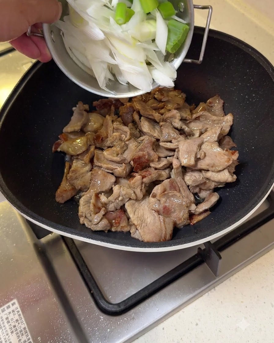 Chopped large scallion sections being added to stir-fried pork in a pan.
