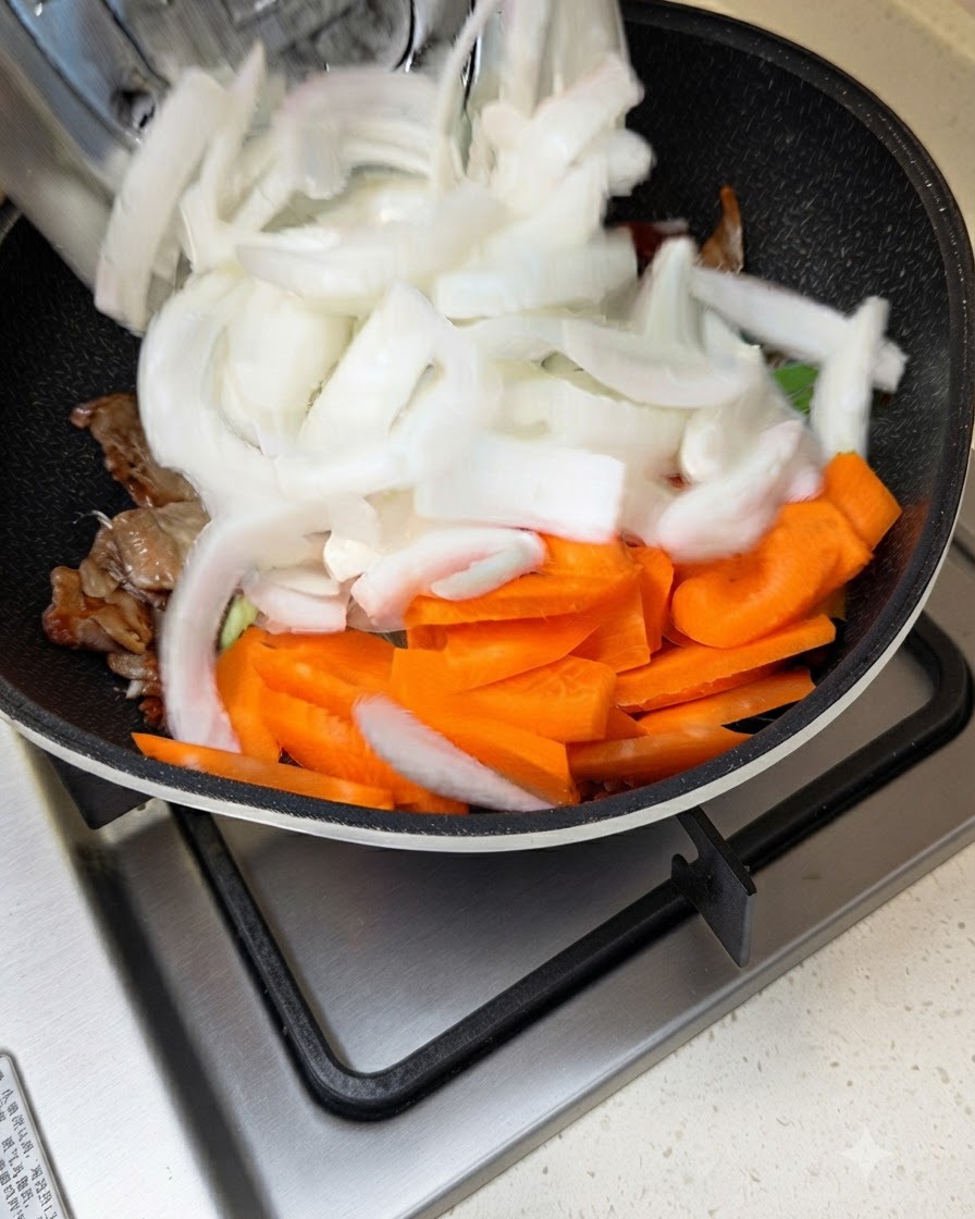 Sliced white onions and carrots being added into a pan with stir-frying meat and sauce.