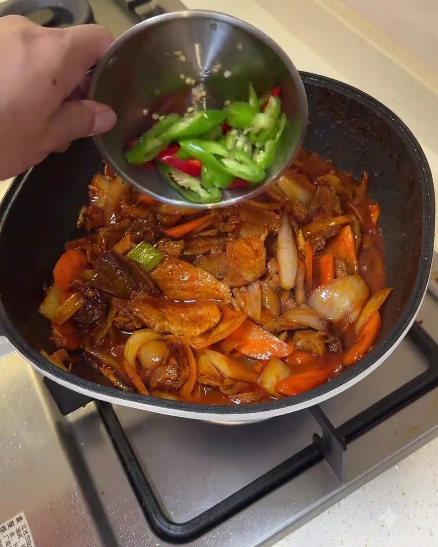 Green and red chili pepper slices being poured from a small bowl into a pan of stir-fried pork and vegetables.