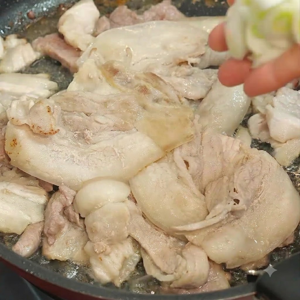 A hand dropping chopped green onions into a pan filled with frying pork slices.