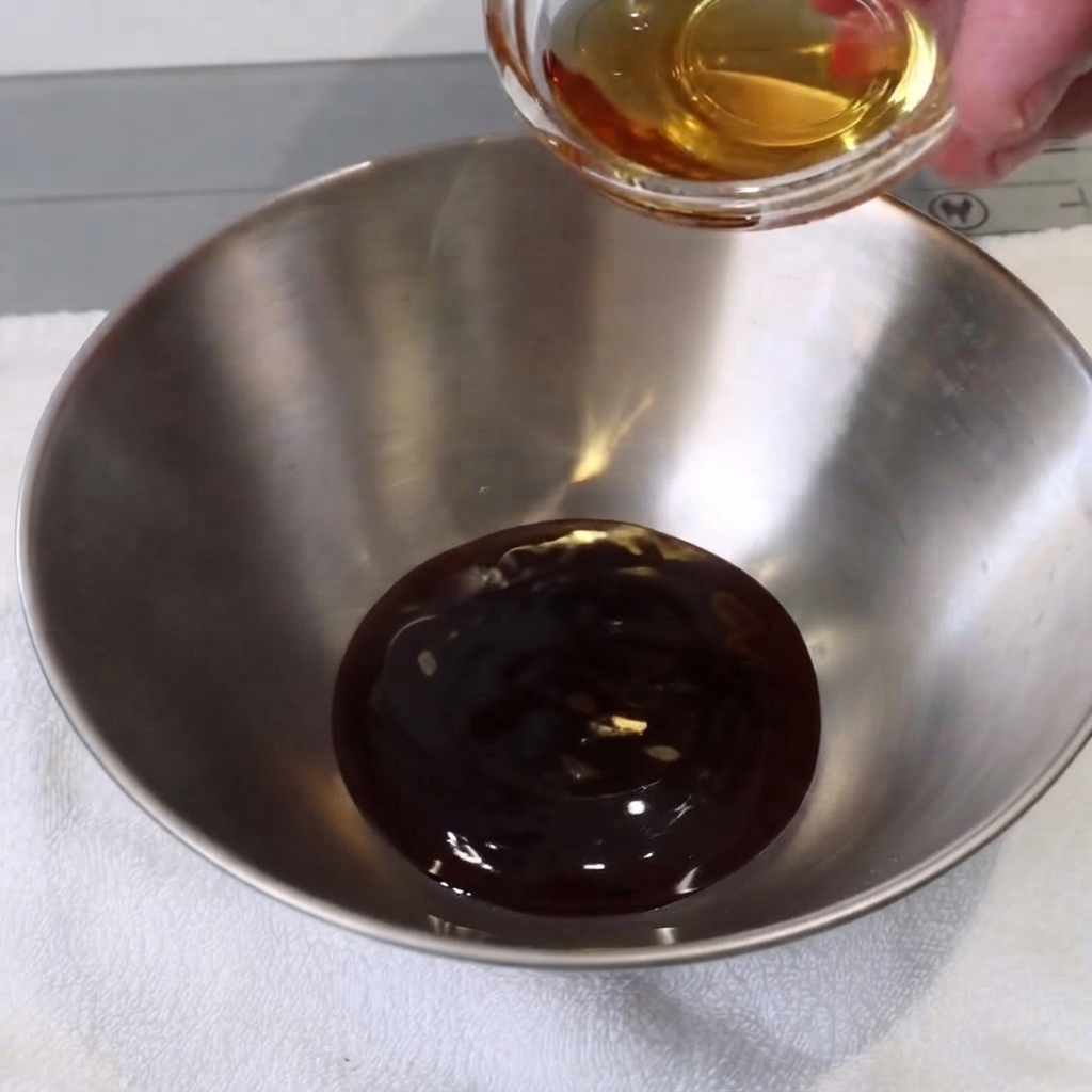 A small metal bowl with liquid ingredients being stirred with a spoon on a white towel.