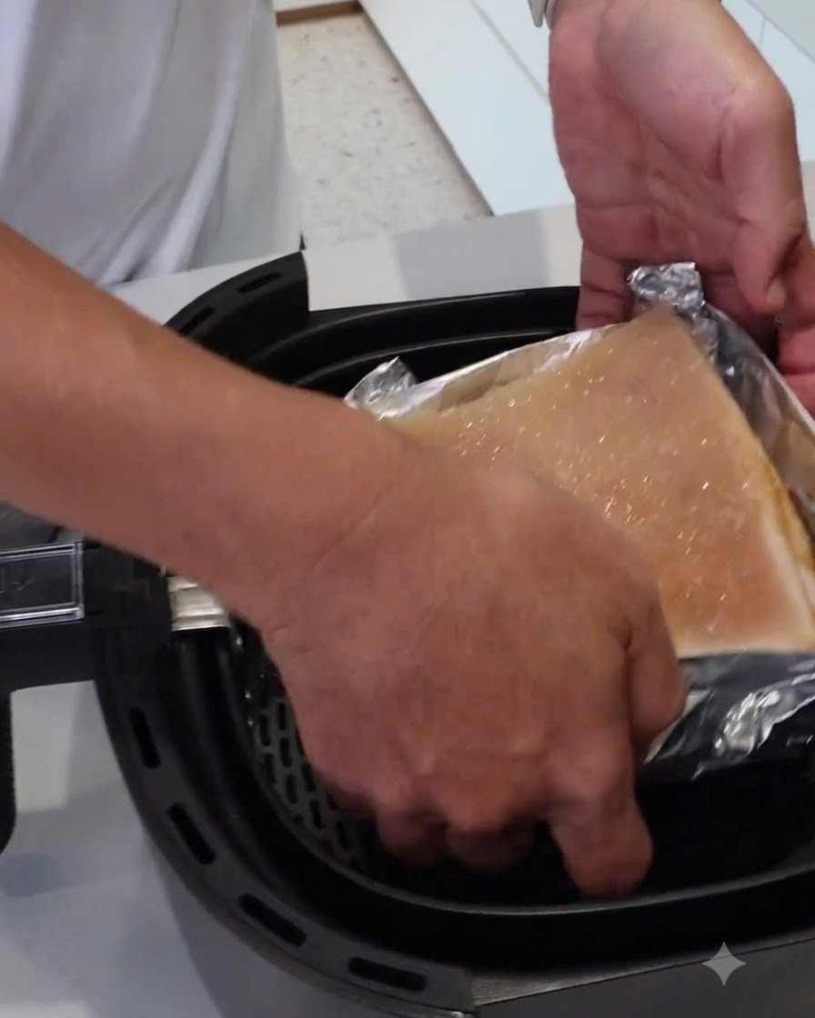 A pair of hands lowering a foil-wrapped block of meat into an air fryer basket.