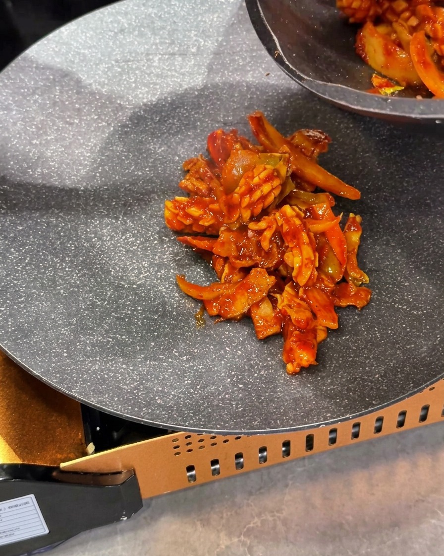 The cooked red squid and pork stir-fry mixture being placed in the center of a portable gas stove skillet.