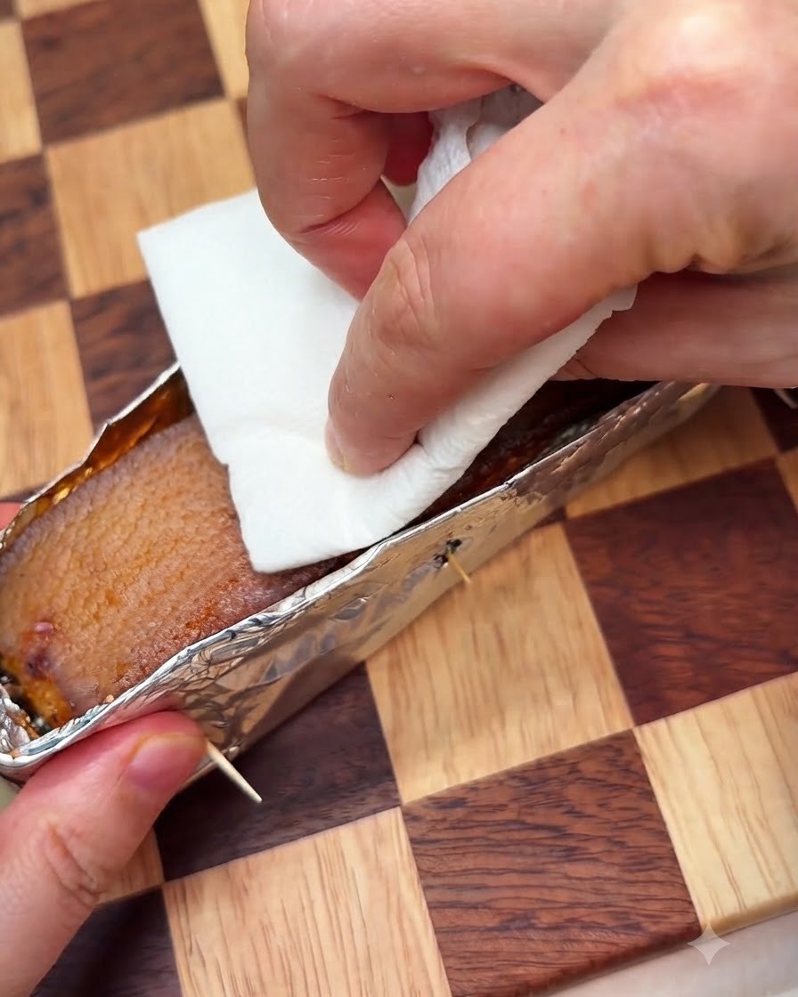 A paper towel being pressed onto the pork skin inside a foil tray to absorb moisture while being pricked.