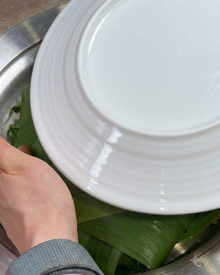 Hands folding the edges of green Zong leaves inward over the ingredients, covered with a white plate.