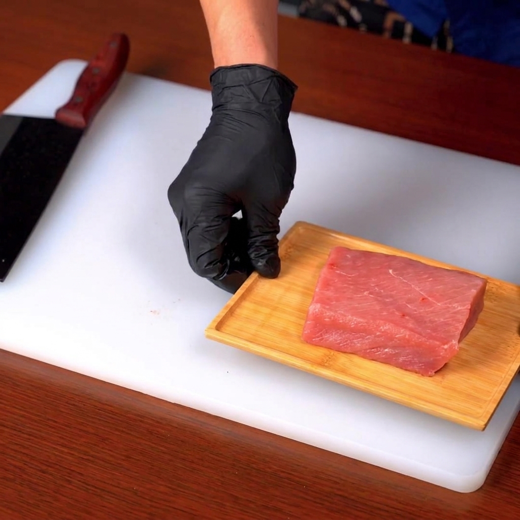 A cut of raw pork loin being sliced on a wooden board atop a white cutting surface.