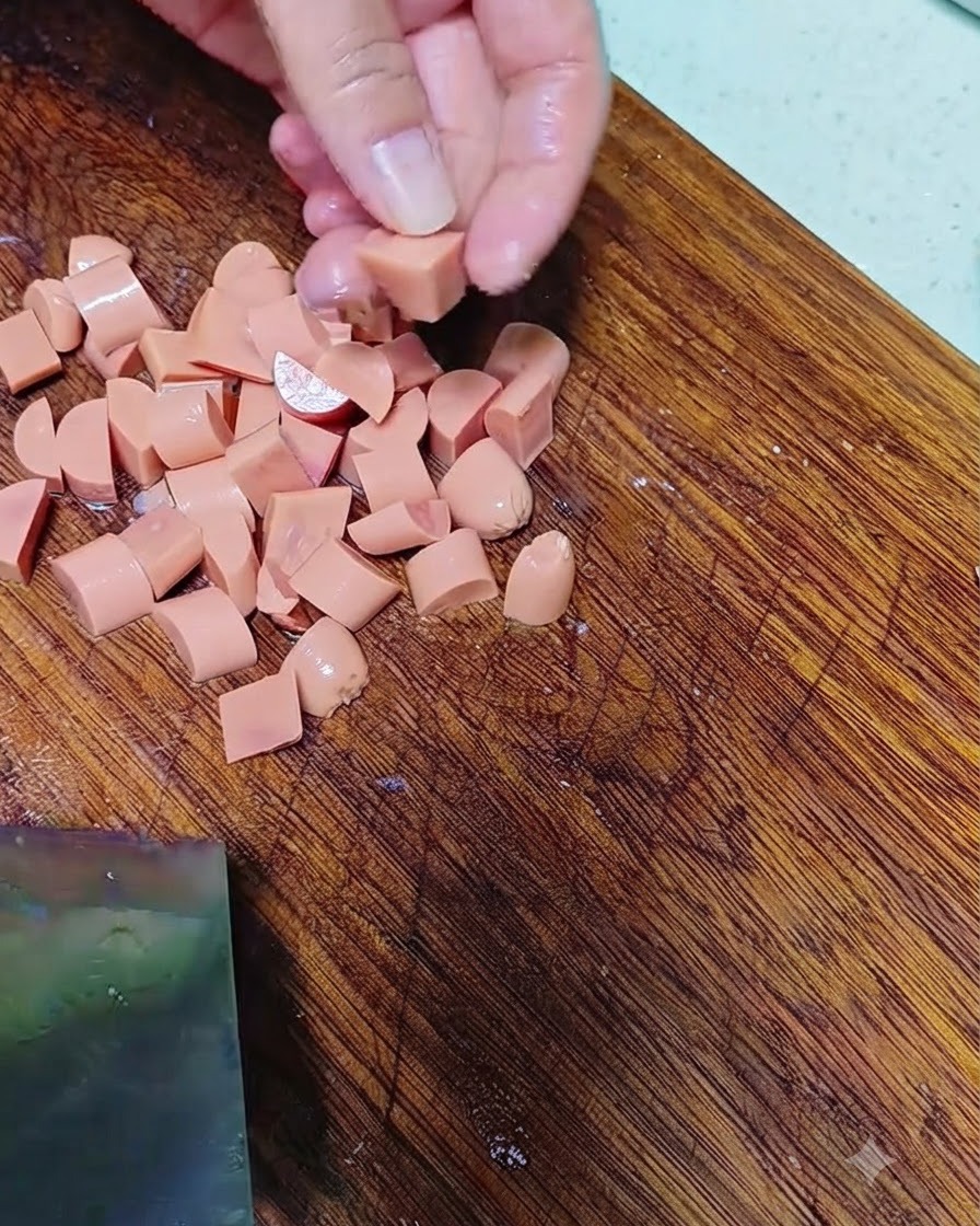 Hands cutting sausage into small pieces on a wooden cutting board.