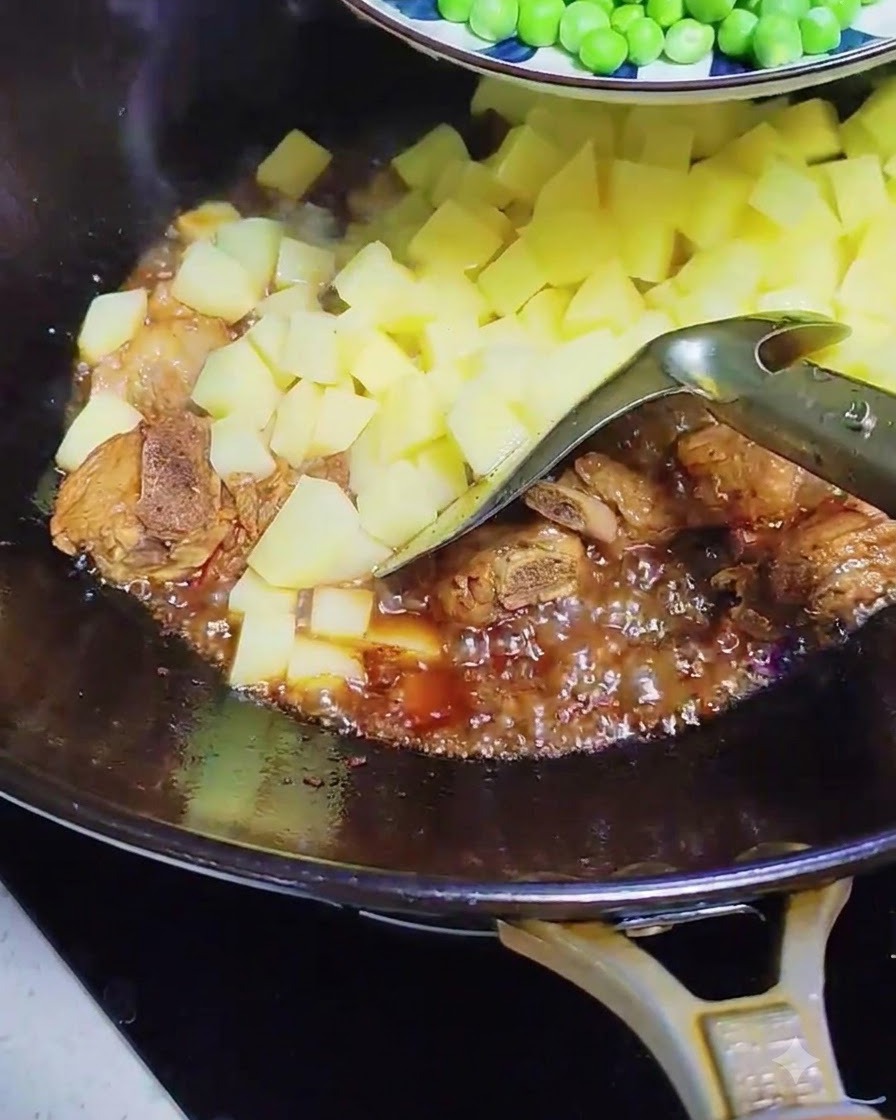 Green peas and sausage slices being added into the wok with bubbling pork ribs and potatoes.