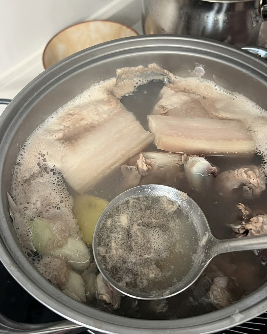 A slotted spoon skimming grey foam and impurities from boiling water in a pot containing pork ribs.