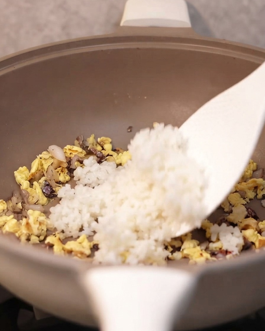 A spatula adding cooked white rice into the pan with the seasoned ingredients.