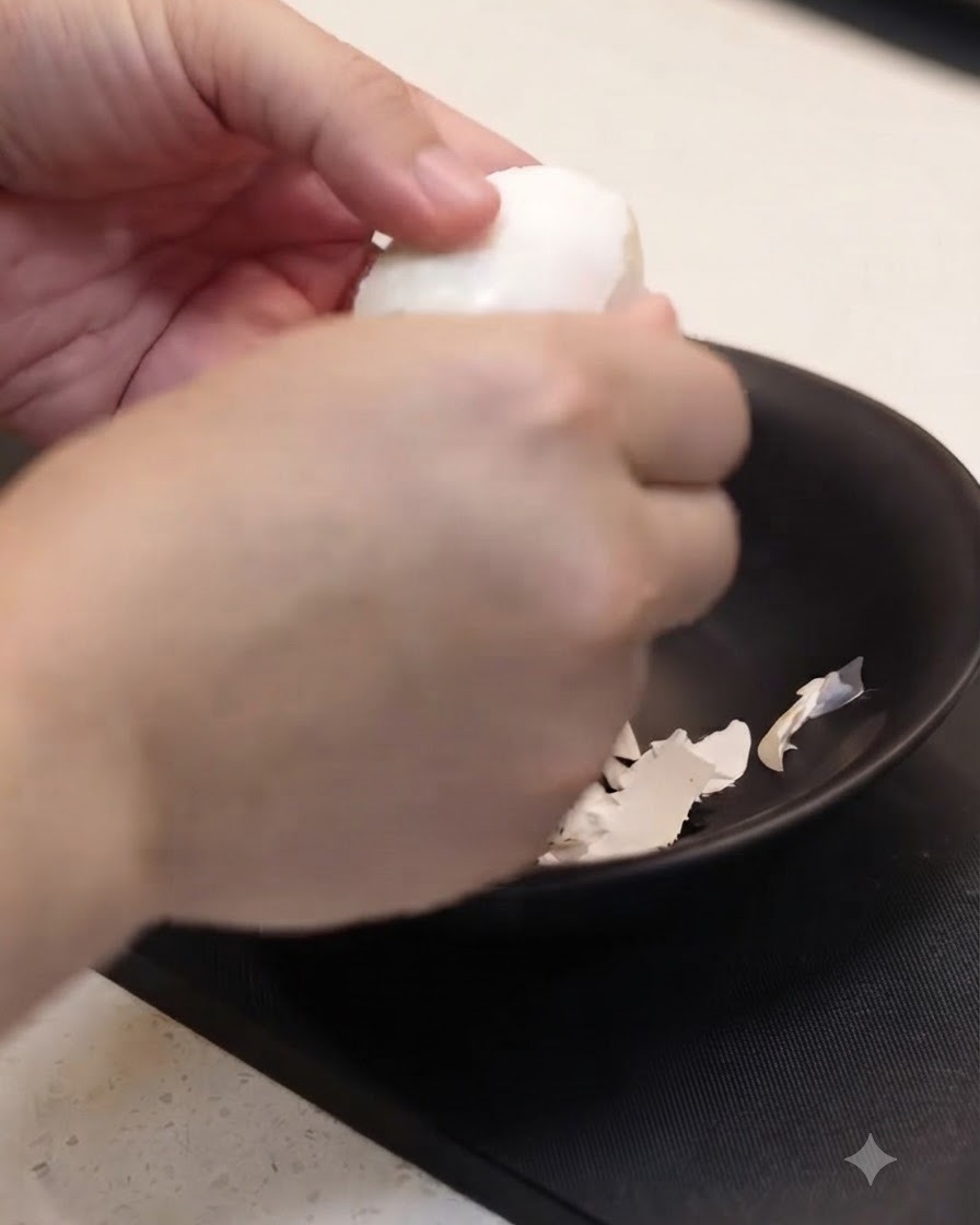 A cooked egg being carefully peeled by hand over a black bowl.