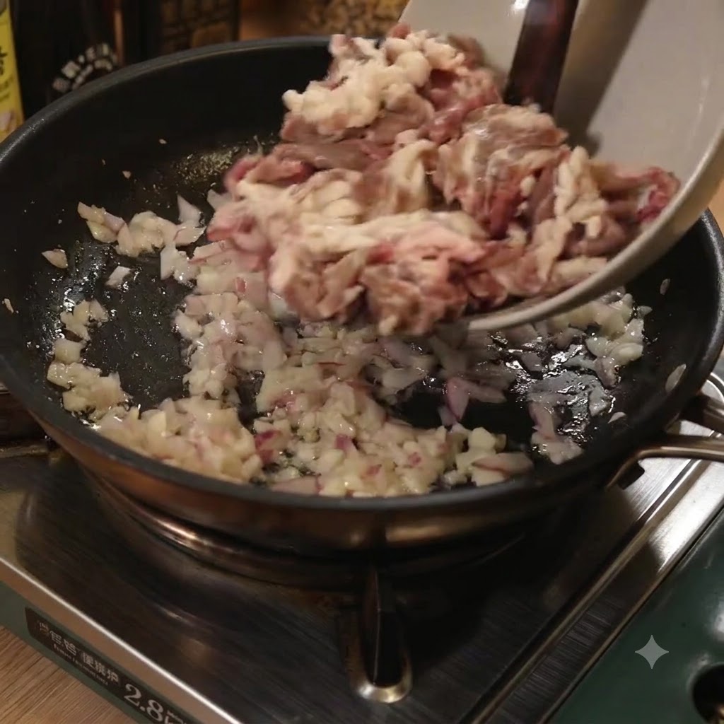 Thin strips of beef being added from a bowl into a frying pan with sautéed onions and garlic.