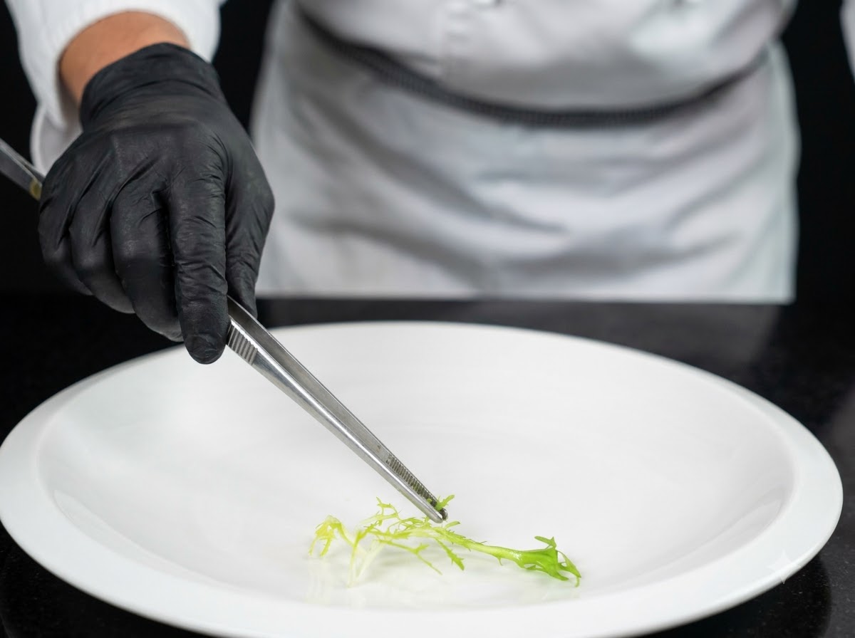 A chef wearing black gloves using tongs to place fresh green vegetable garnish onto the center of a large white plate.