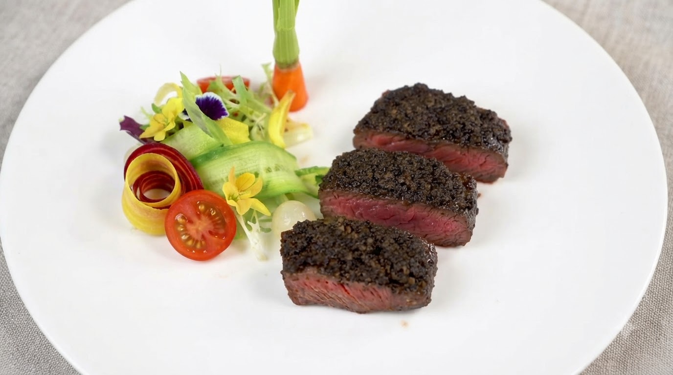 Three thick slices of medium-rare steak topped with rich black truffle paste, served alongside fresh vegetable garnishes.