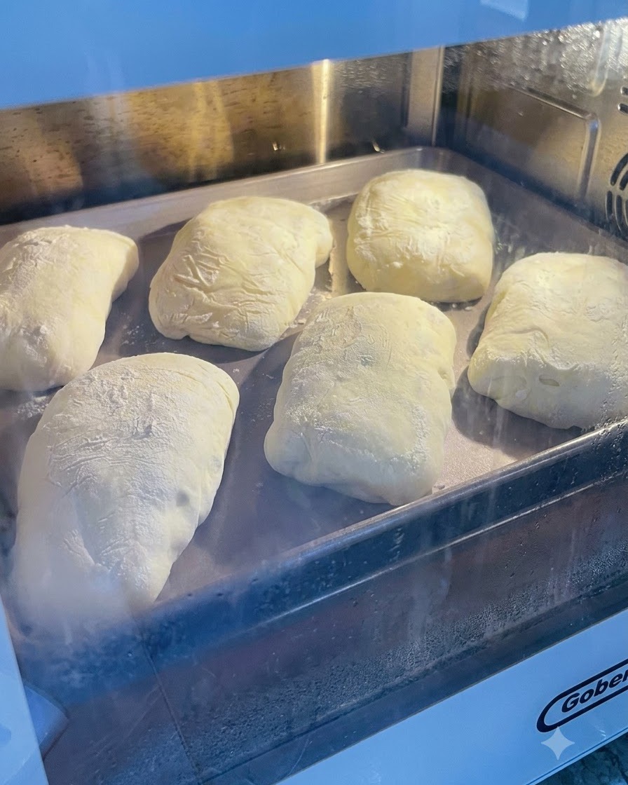 Fermented dough dusted with flour sitting on a metal baking tray inside an oven or proofing area.