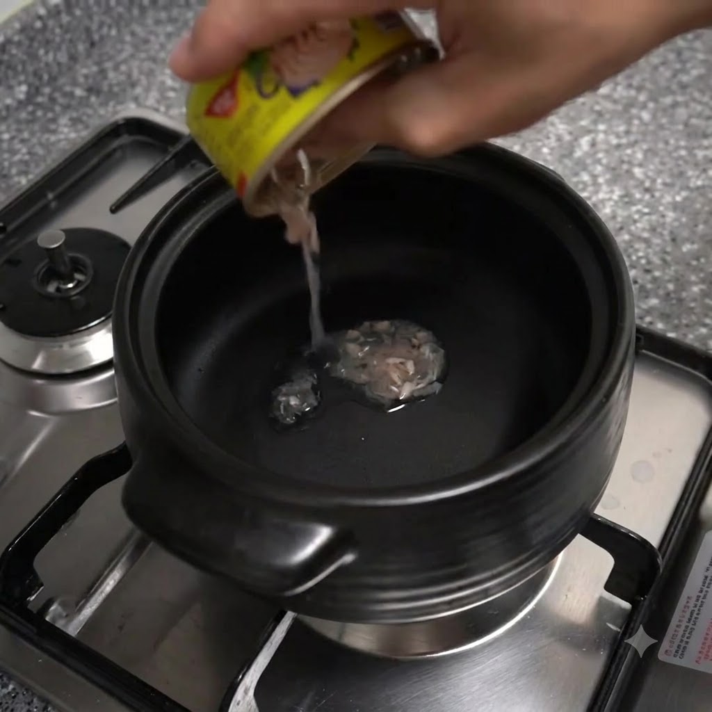 Oil being poured from a small yellow-labeled tuna can into a black clay pot resting on a stove burner.