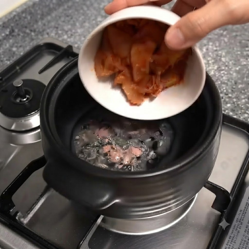 A white bowl tilting and dropping chopped red kimchi pieces into the black clay pot.