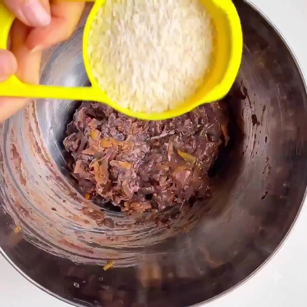 Oat flour being poured from a yellow measuring cup into a metal mixing bowl with mashed beans and vegetables.