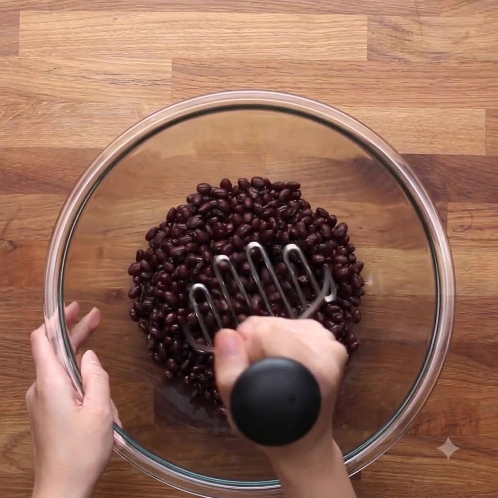 A metal potato masher pressing down into a bowl of black beans to mash them.