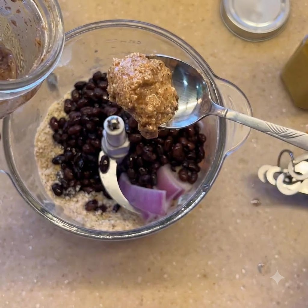 A spoonful of almond butter being held over the blender containing black beans and red onioncite: 1.