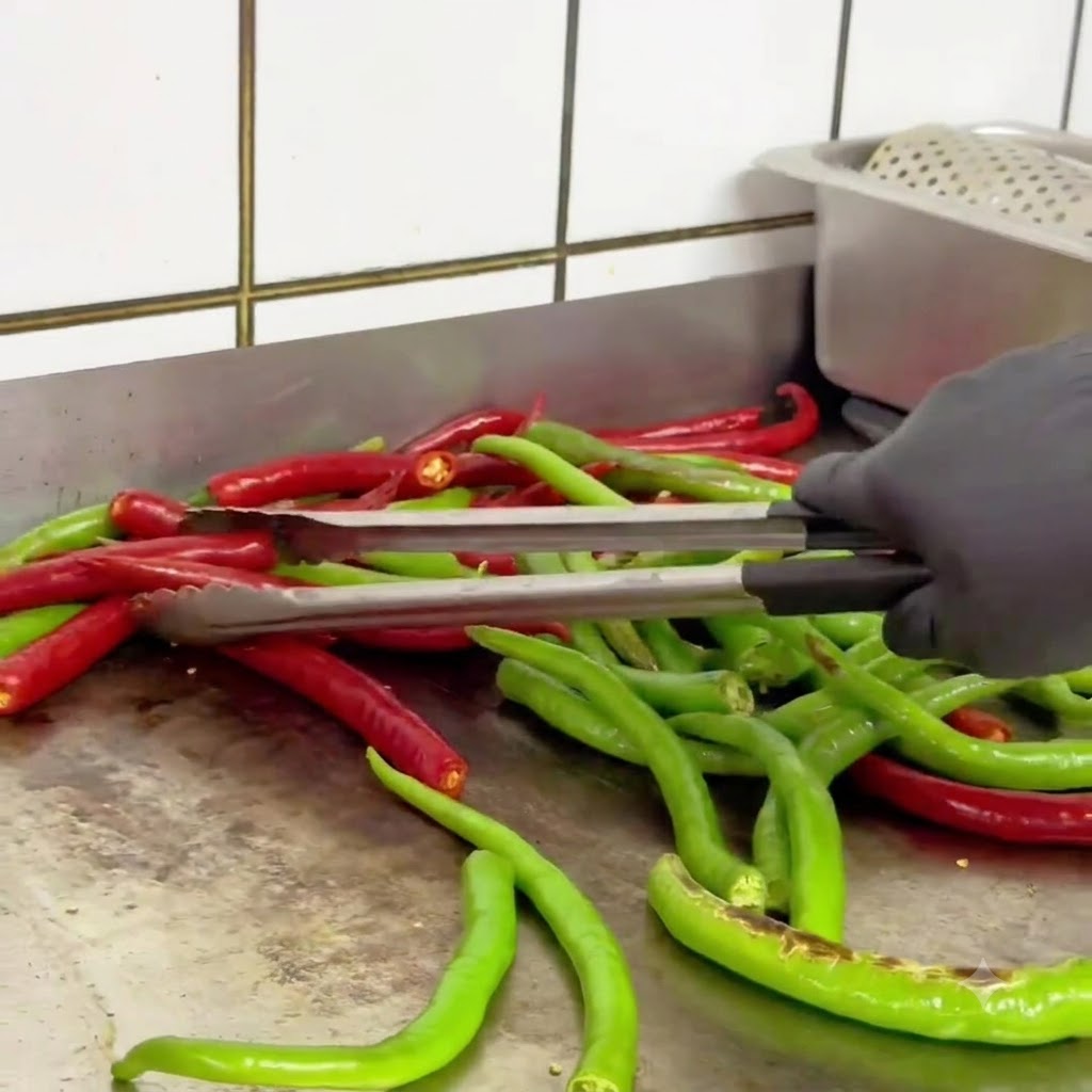 Green and red chili peppers being turned with tongs in a hot pan while charring.